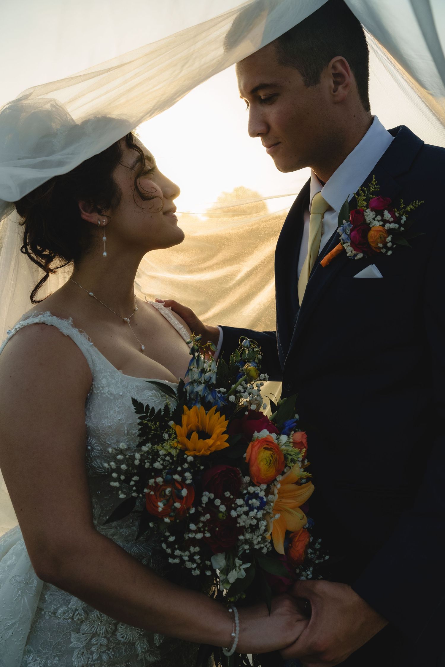 Intimate wedding couple moment with sunflower bouquet and flowing veil at sunset.