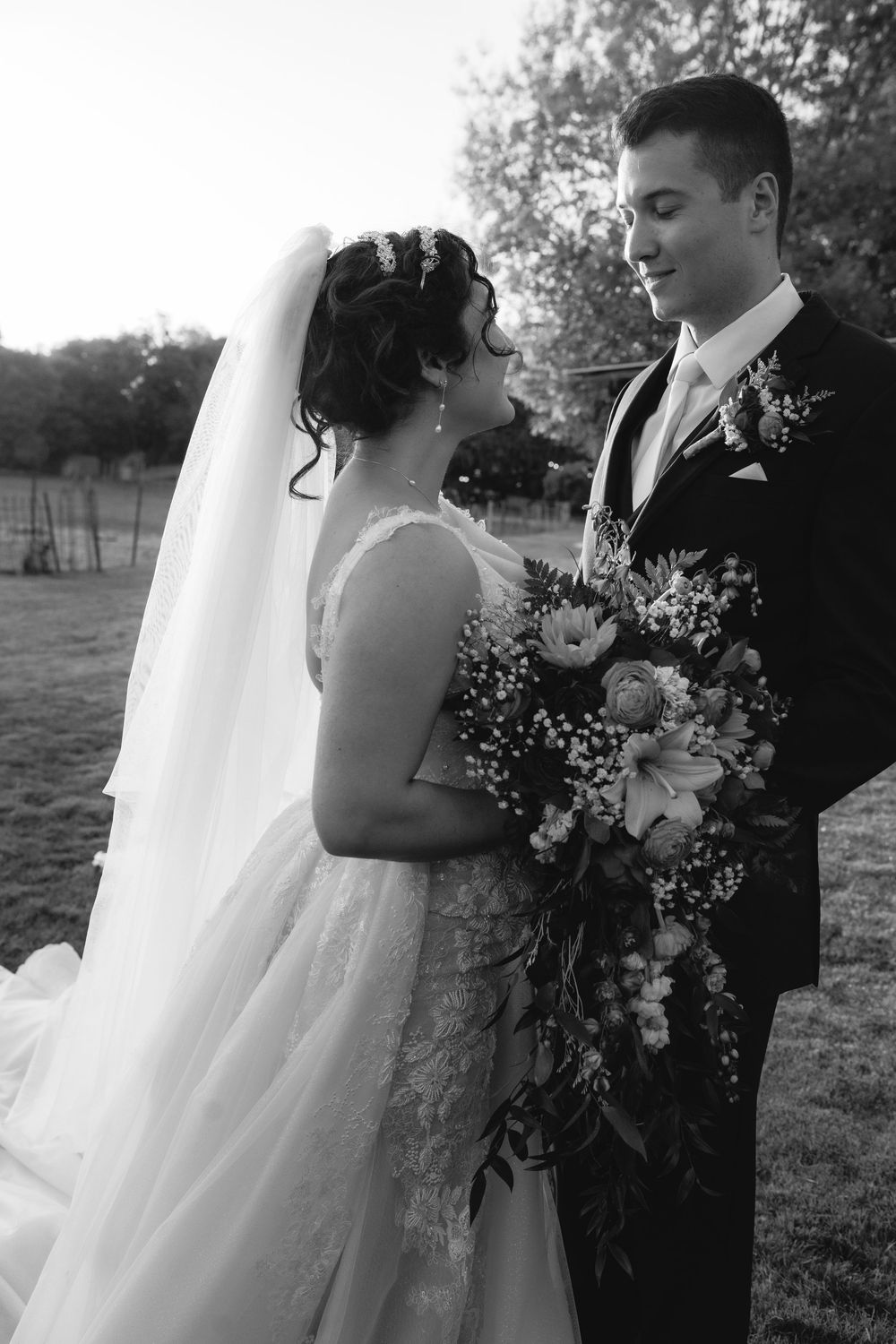 Black and white wedding portrait with flowing veil and floral bouquet in outdoor setting.