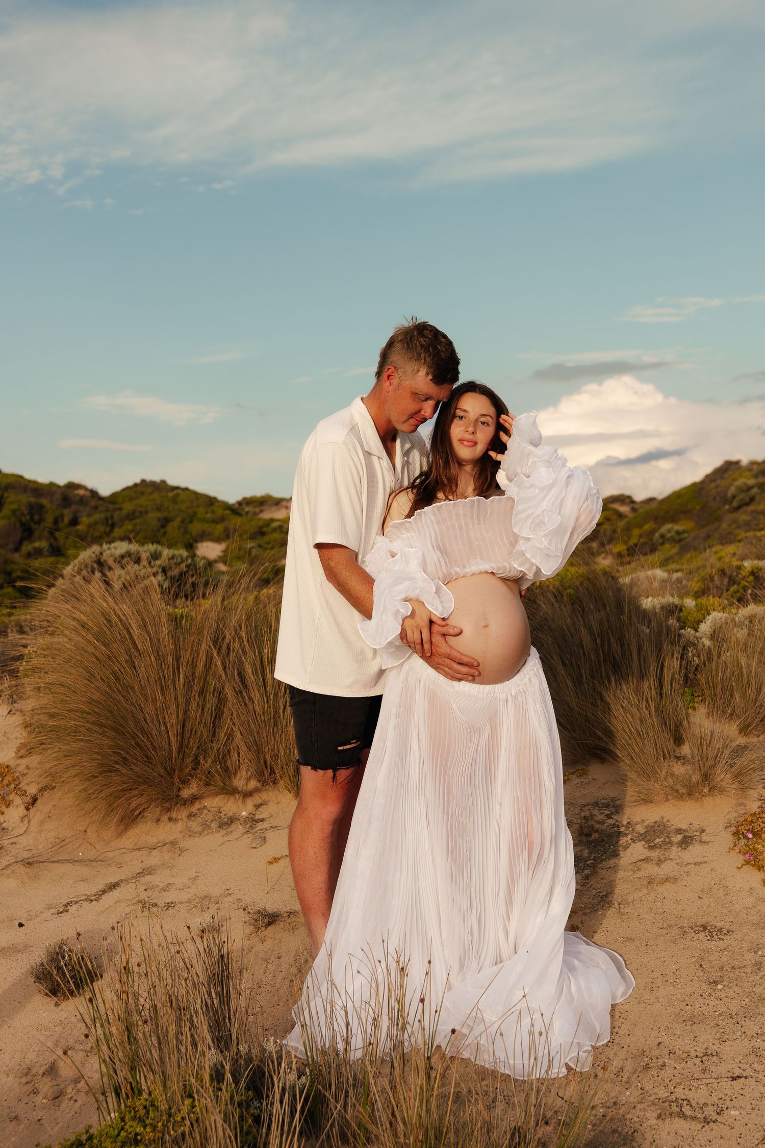 A person in a flowing white off-shoulder dress poses on a sandy beach against dramatic rocky cliffs at sunset.