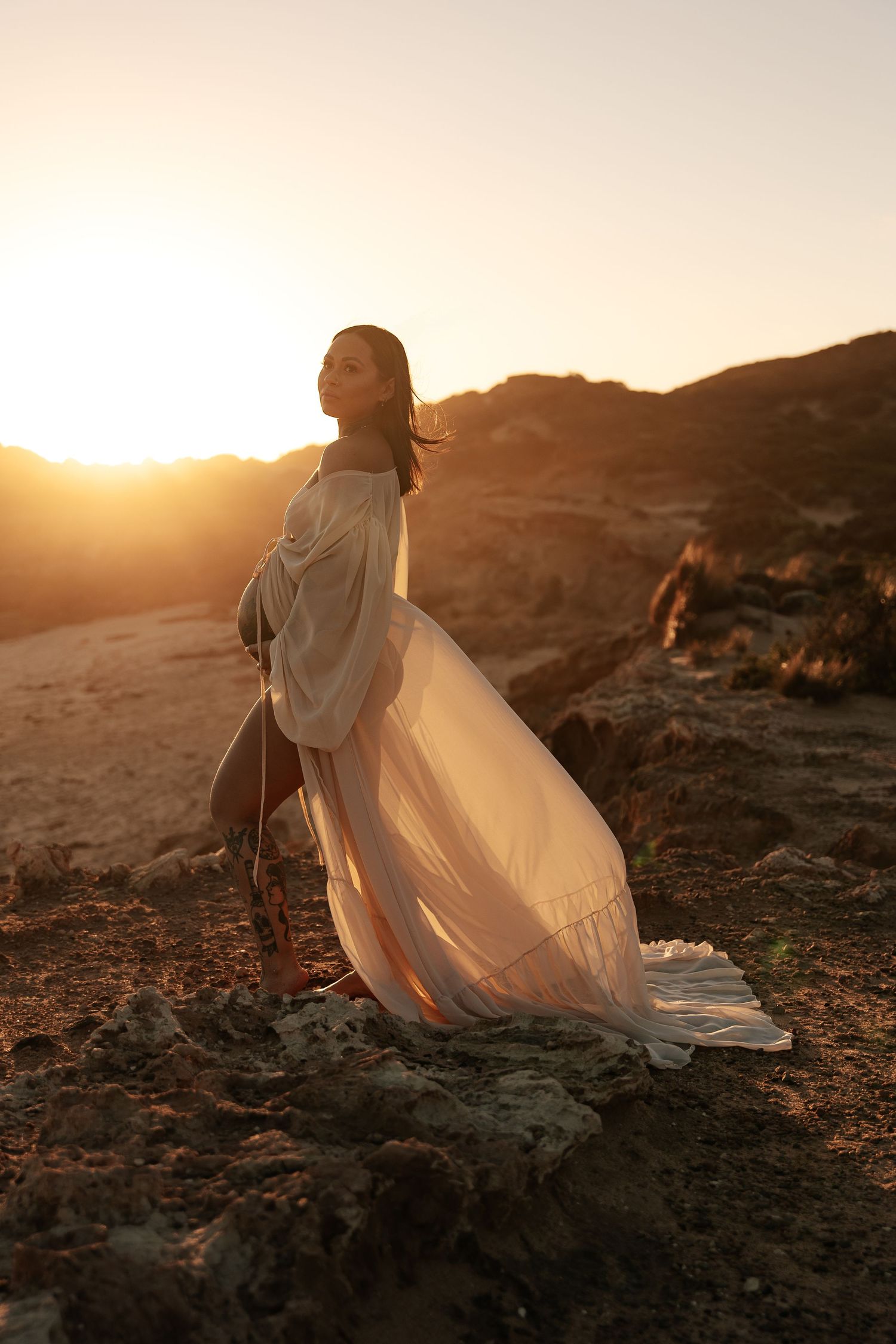 A person in a flowing white off-shoulder dress poses on a sandy beach against dramatic rocky cliffs at sunset.
