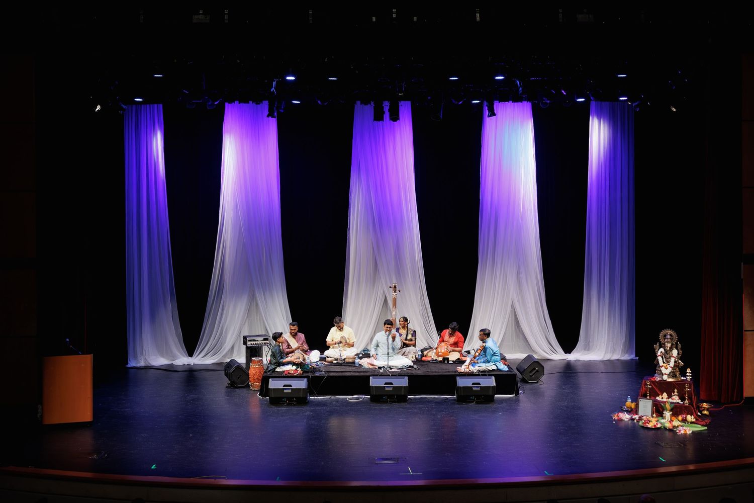 Musicians perform traditional Indian classical music on stage with dramatic purple and white draped backdrop lighting.