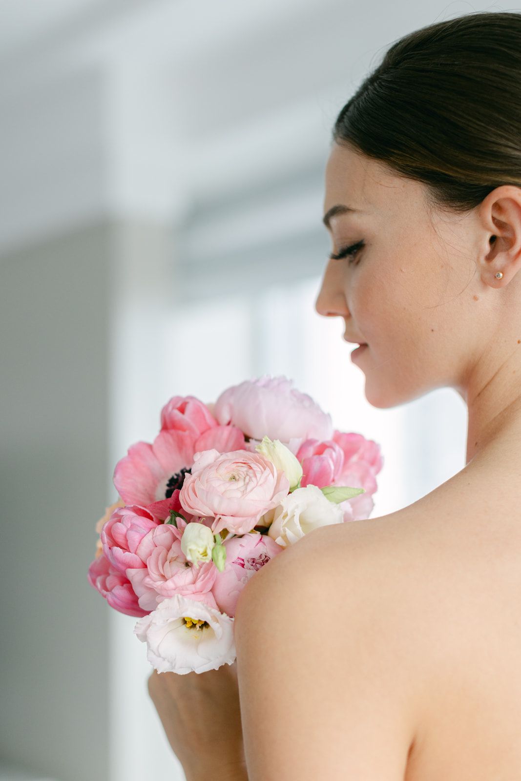 White lace bridal heels sit beside pink and peach roses in a vase on tulle fabric by a bright window.
