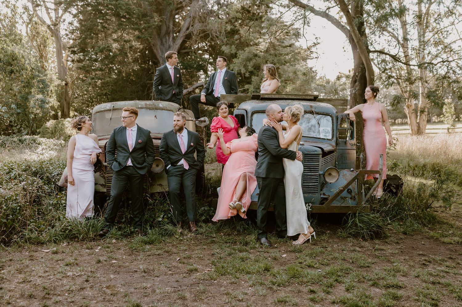 A vintage black car serves as a rustic backdrop for a wedding party posing in formal attire in a wooded setting.