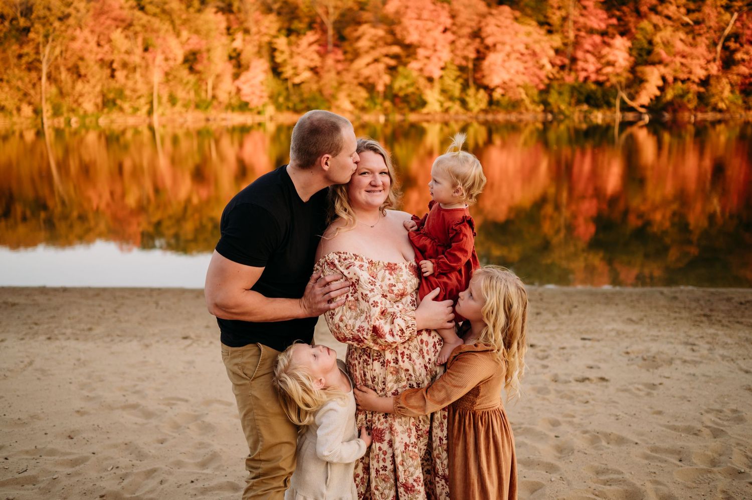 A family embraces on a sandy beach during golden hour with autumn foliage reflecting in the water behind them.
