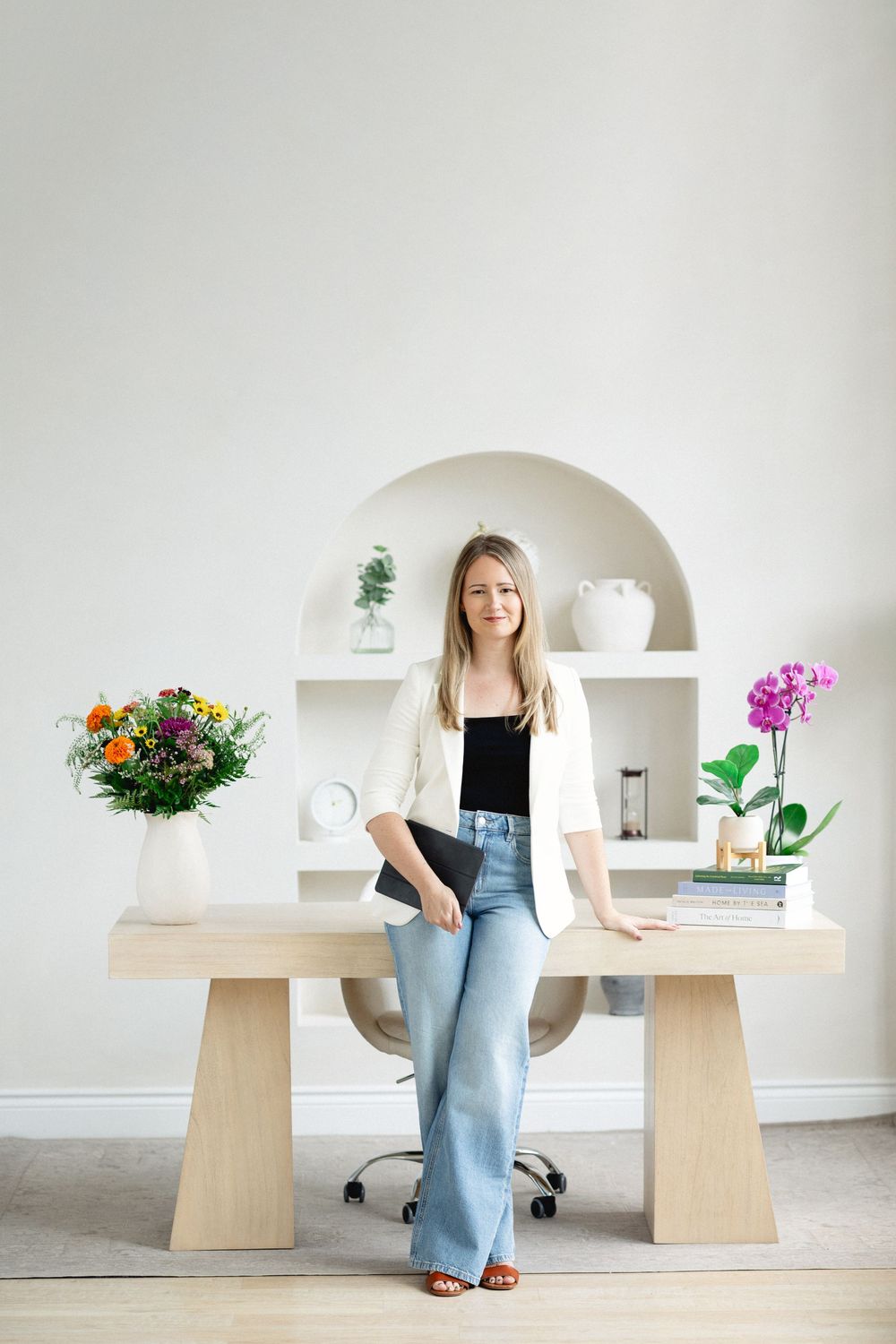 virtual Ontario therapist standing in front of clean minimal desk in light setting with white blazer and blue jeans holding a laptop