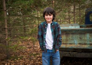 A child in denim and plaid stands near a wooden fence in a wooded outdoor setting.