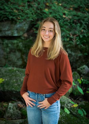 A person in a rust-colored sweater and blue jeans poses outdoors against a green natural background.