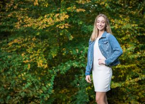 A person in a denim jacket and white dress poses against vibrant autumn foliage in a forest setting.