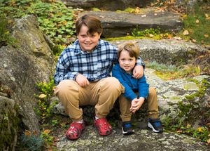 Two young friends sit together smiling on large rocks surrounded by autumn foliage in an outdoor setting.
