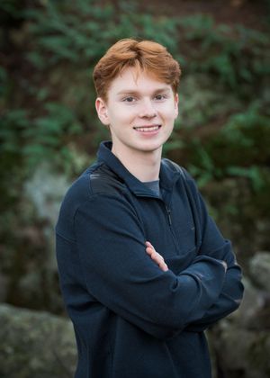 Professional portrait photo of a smiling person in a dark pullover against a blurred outdoor background.