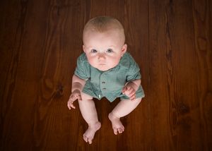 High angle portrait of infant wearing sage green outfit lying on dark wooden floor in studio setting.