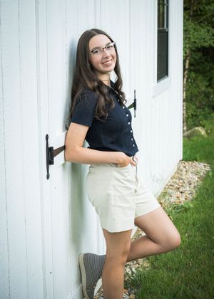 A woman in glasses and navy top leans against a white wall wearing khaki shorts and casual shoes outdoors.