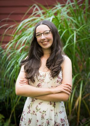 Portrait of someone wearing glasses and a floral dress standing with crossed arms against a foliage background.