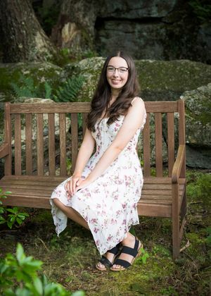 Portrait of someone in a floral summer dress and glasses seated on a wooden bench in a garden setting.