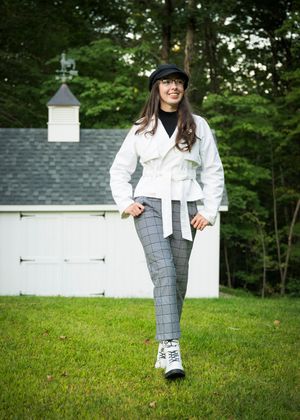 A figure in plaid pants, white jacket and black hat stands casually on grass with a white building in the background.