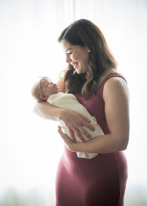 A person in a burgundy sleeveless top holds and gazes lovingly at a swaddled newborn baby in a bright studio setting.