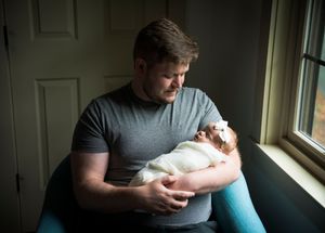 A person in a gray t-shirt sitting in natural window light holds a tiny white infant swaddled in white cloth.