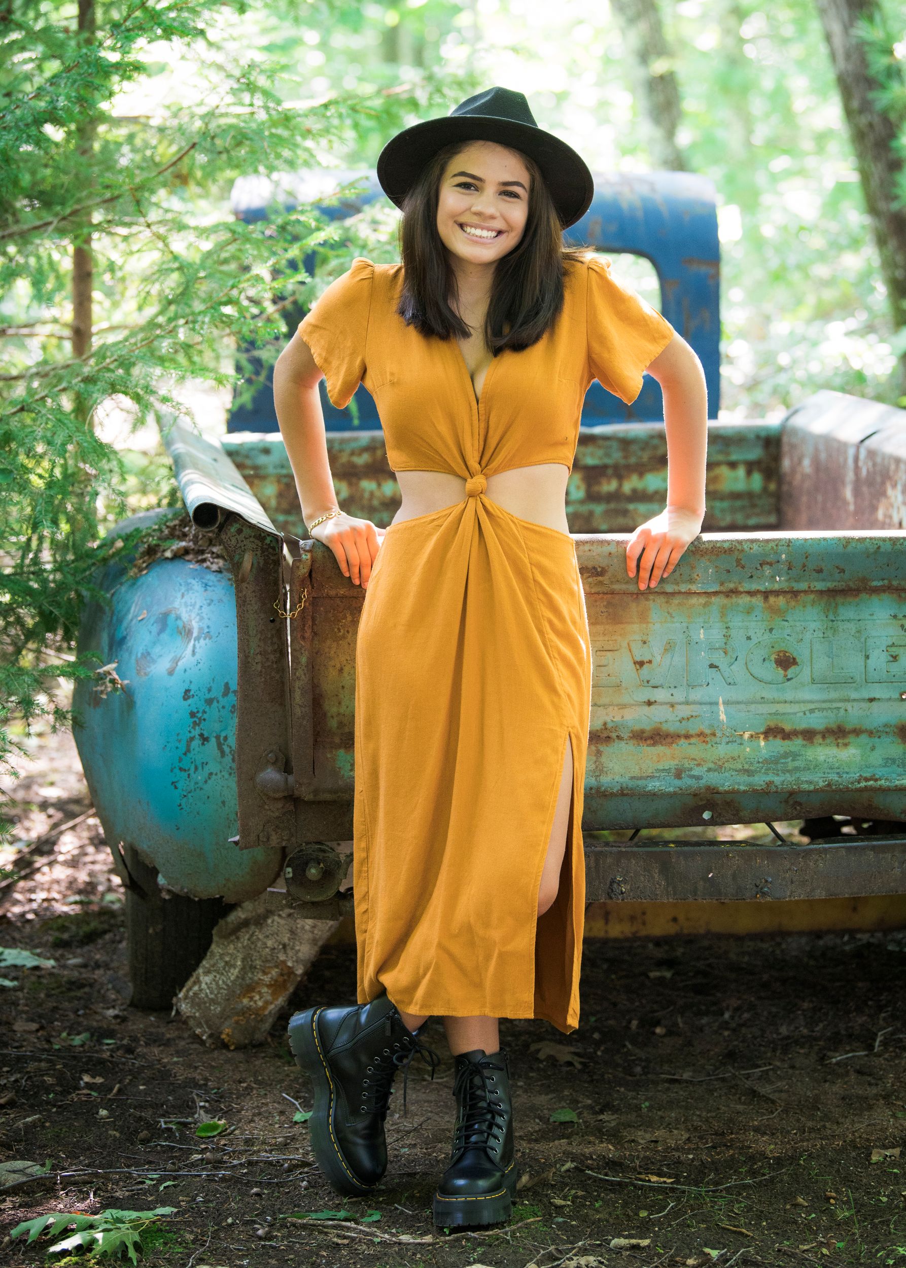 Rustic outdoor portrait featuring a yellow twist-front dress, black hat and combat boots against vintage truck backdrop.