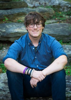 Someone in a blue polka dot shirt and glasses sits on rocky terrain wearing bracelets while smiling at the camera.