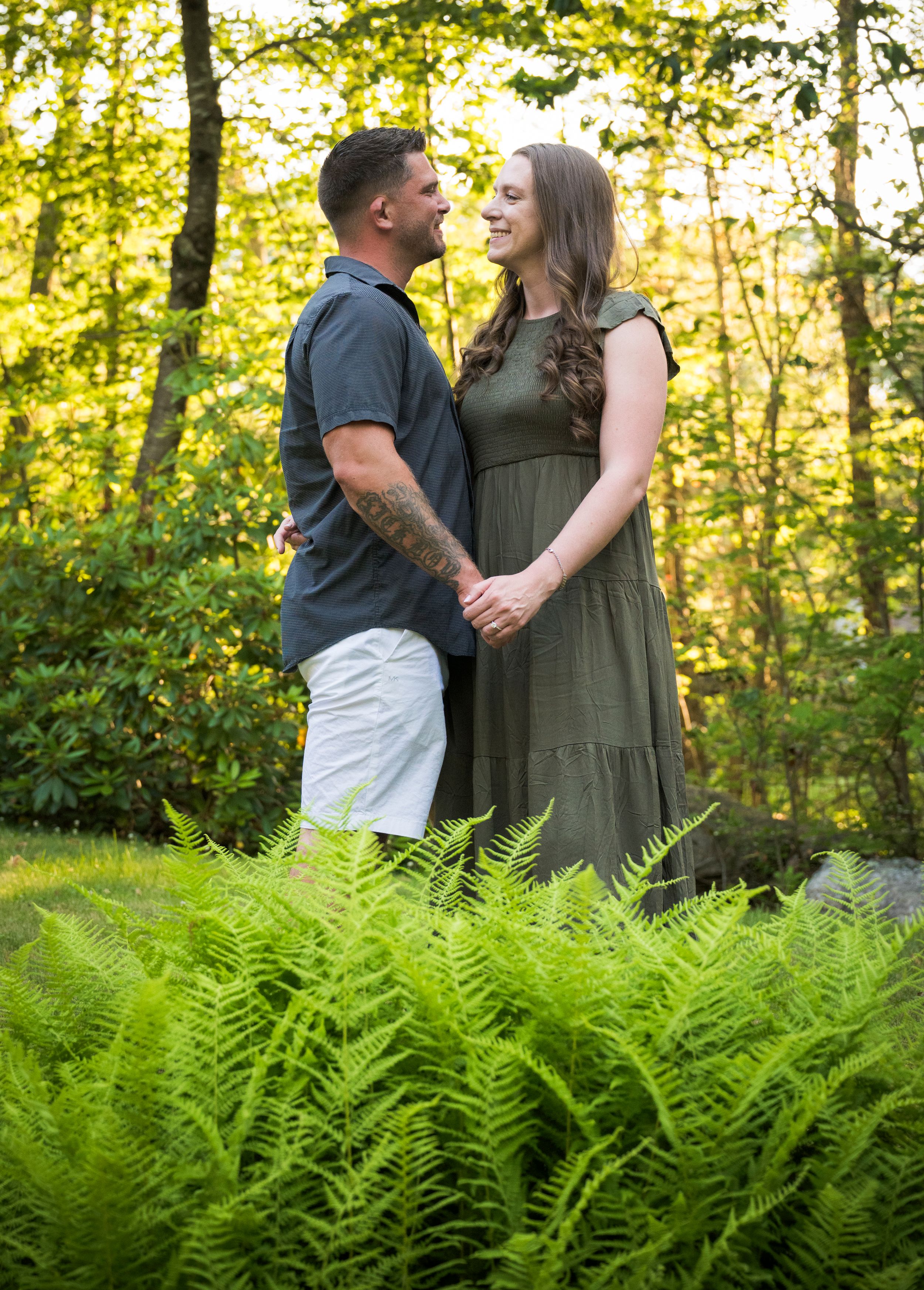 A couple shares a romantic moment in a forest clearing with lush green ferns in the foreground.