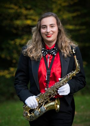 Concert performer in black suit and red tie holds a saxophone while standing against a green outdoor backdrop.