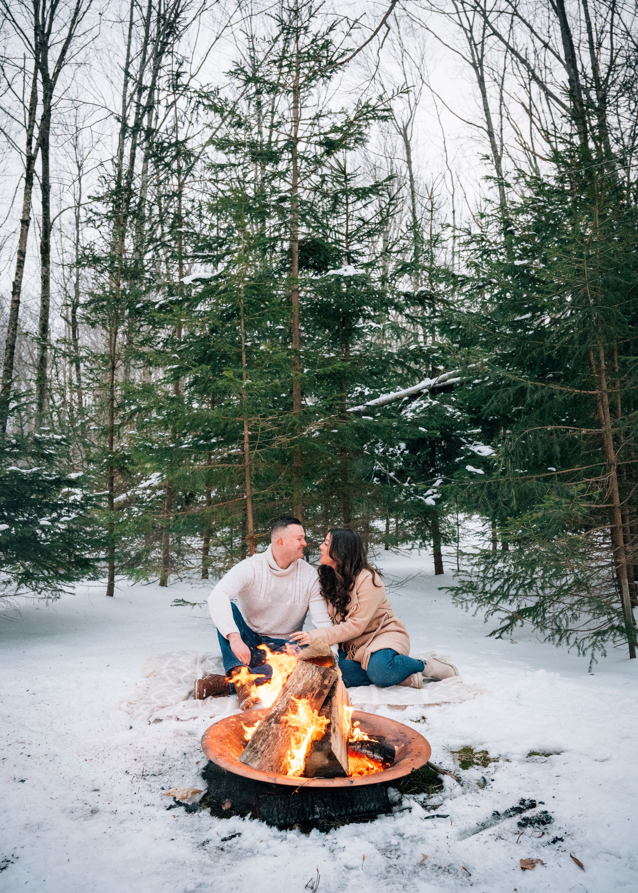 A couple sits on a blanket near a glowing campfire in a snowy forest setting with evergreen trees in the background.