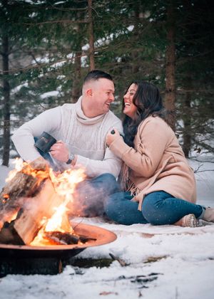 A couple shares a romantic moment lying in the snow near a cozy campfire in a winter forest setting.