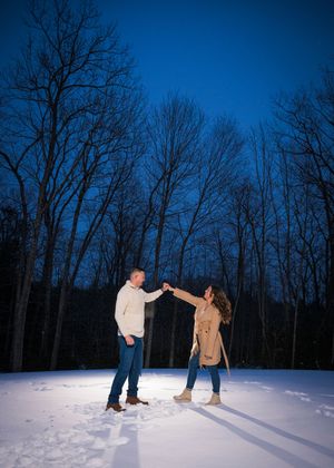 Two people dance together in the snow at dusk with bare winter trees silhouetted against a deep blue sky.