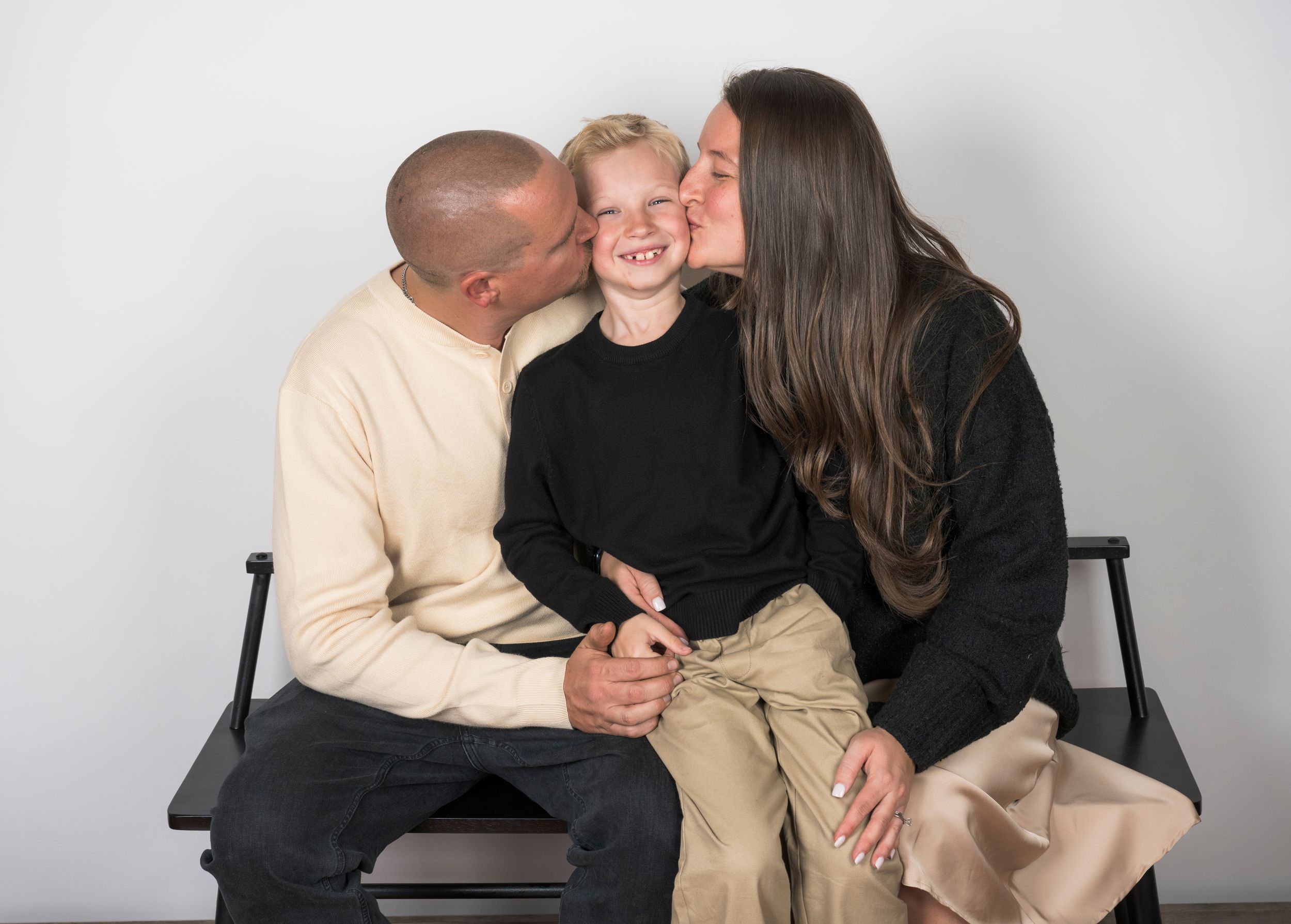 A family of three sits together on a black bench sharing a sweet moment against a light gray background.