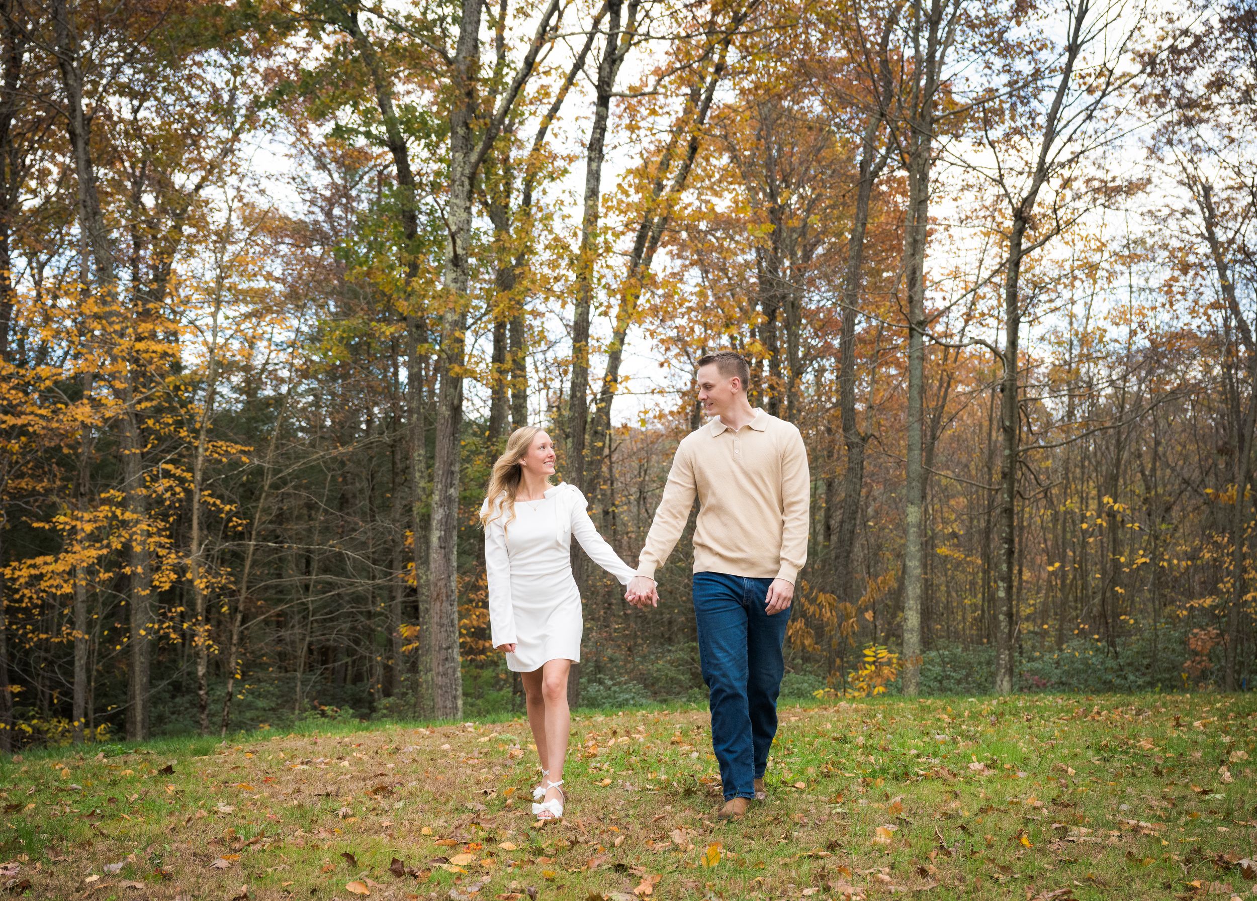 A couple walks through an autumn forest with golden leaves falling from the trees during a romantic photoshoot.