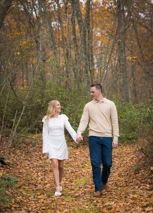 A couple walks hand in hand through a forest path covered in fallen autumn leaves on a crisp fall day.