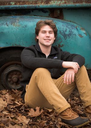 A person wearing a black sweater sits against a vintage turquoise vehicle, surrounded by fallen autumn leaves.