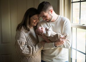 A couple in cozy sweaters lovingly cradles their newborn baby near a sunlit window.