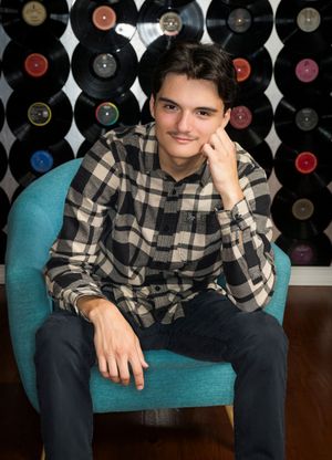 Young person in plaid shirt sits casually on turquoise chair against wall decorated with vintage vinyl records.