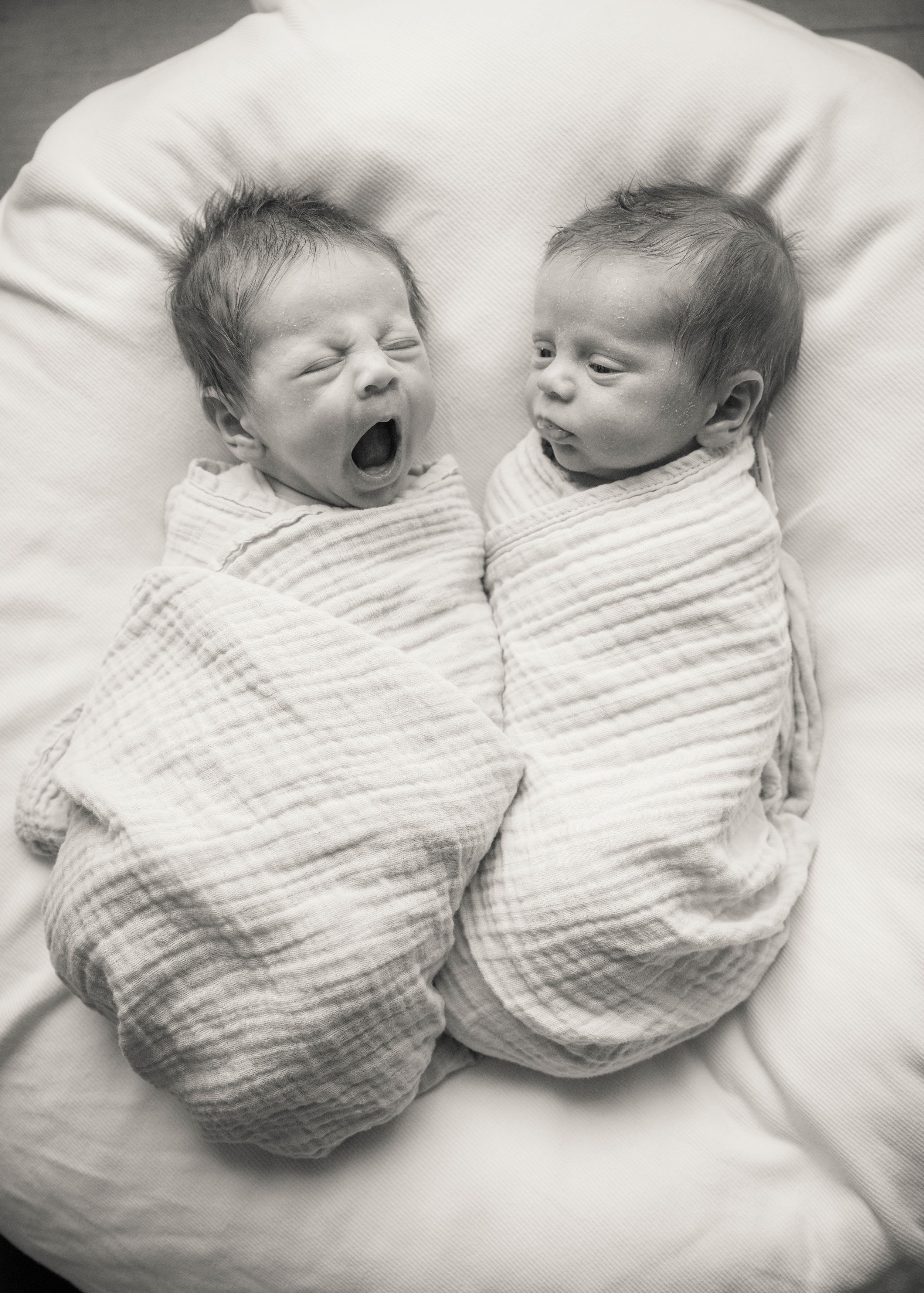 Black and white photo of twin babies swaddled in blankets, one yawning while the other appears peaceful and sleepy.