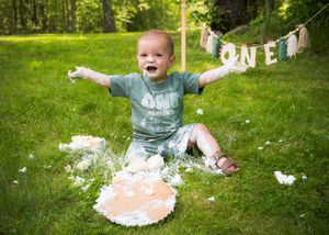 A smiling joyful figure sits in grass during a cake smash photoshoot with white frosting everywhere against a green backdrop.