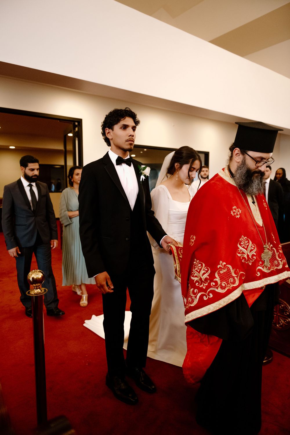 Wedding ceremony procession inside a church with an Orthodox priest in red vestments leading on a red carpet.