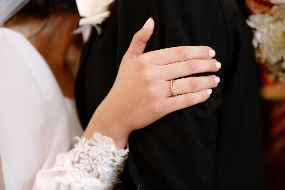 A hand with a wedding ring and lace sleeve detail rests against a black suit jacket at a formal ceremony.