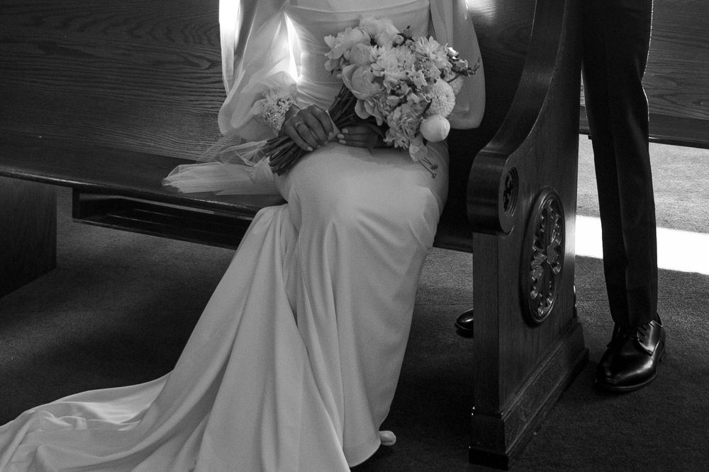 Black and white photograph of an elegant wedding dress with a flowing train and white bouquet beside a dark vehicle.