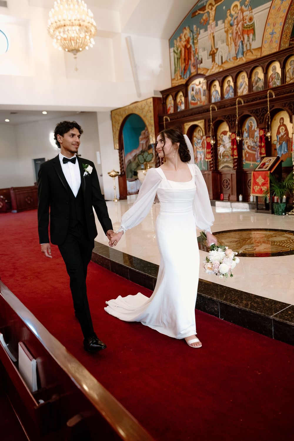 A wedding ceremony in an ornate church with red carpet, featuring a couple walking past an iconostasis with religious art.