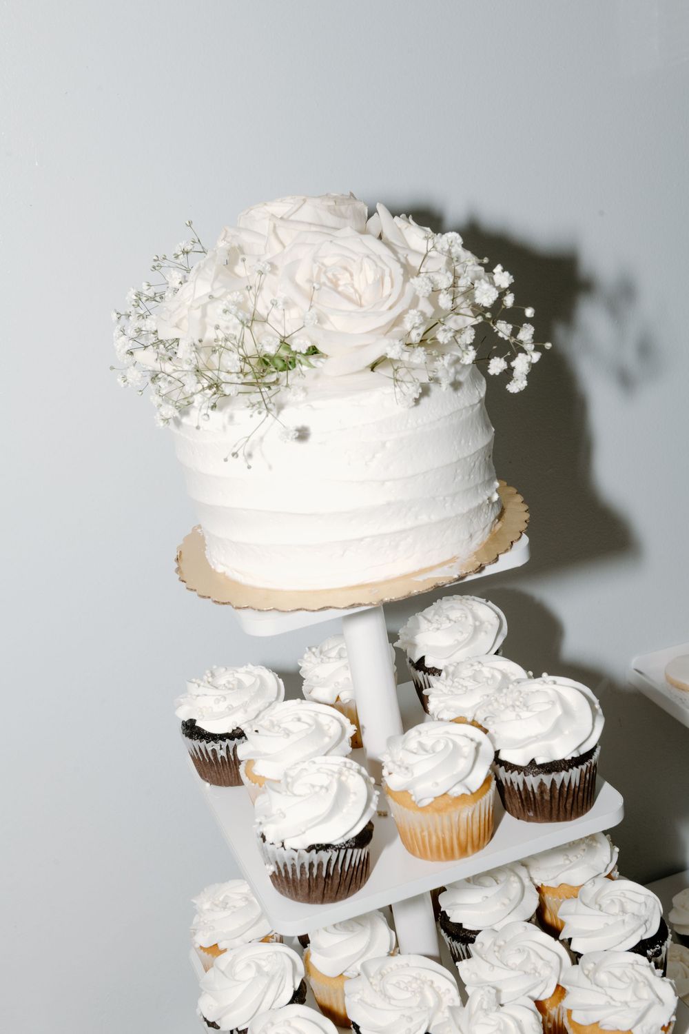 White wedding cake with roses and baby's breath surrounded by decorated cupcakes on tiered display.