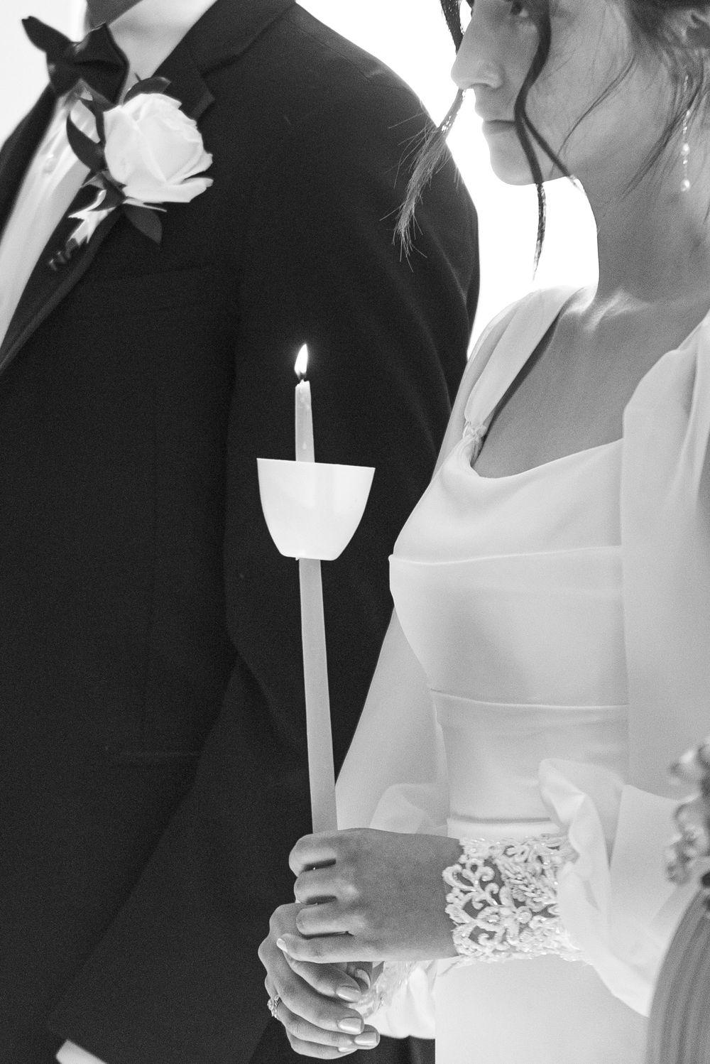 A black and white wedding photo showing a groom in a tux with boutonniere and bride in lace holding a white candle.