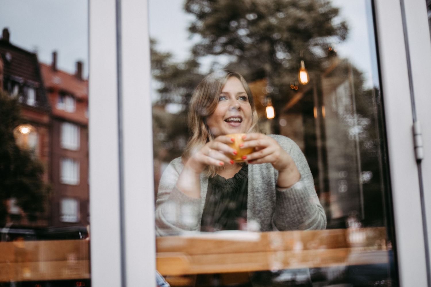 Eine Frau schaut aus lächelnd aus dem Fenster eines Cafès, sie hält eine Tasse in den Händen.