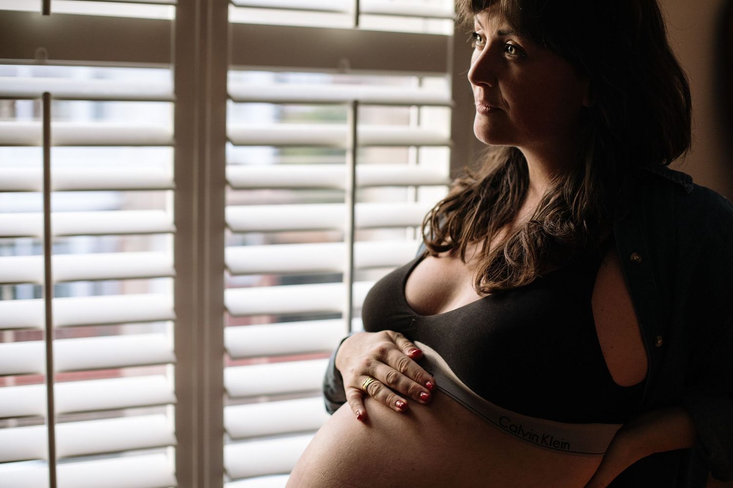 Silhouetted against window blinds, a pregnant woman in black clothing gently cradles her baby bump.