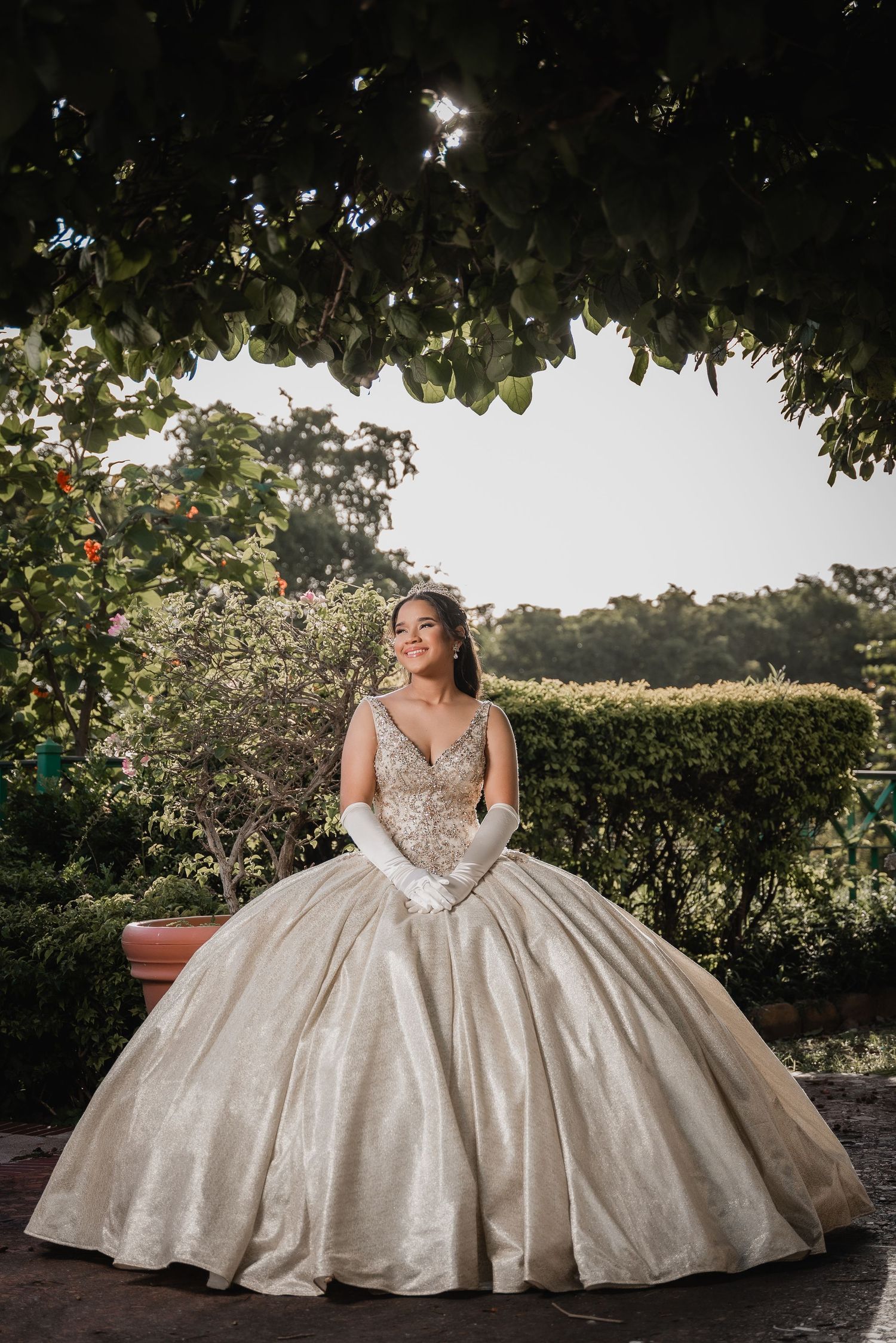 Elegant ballgown with intricate beading and full skirt captured in a garden setting at golden hour beneath overhanging foliage.