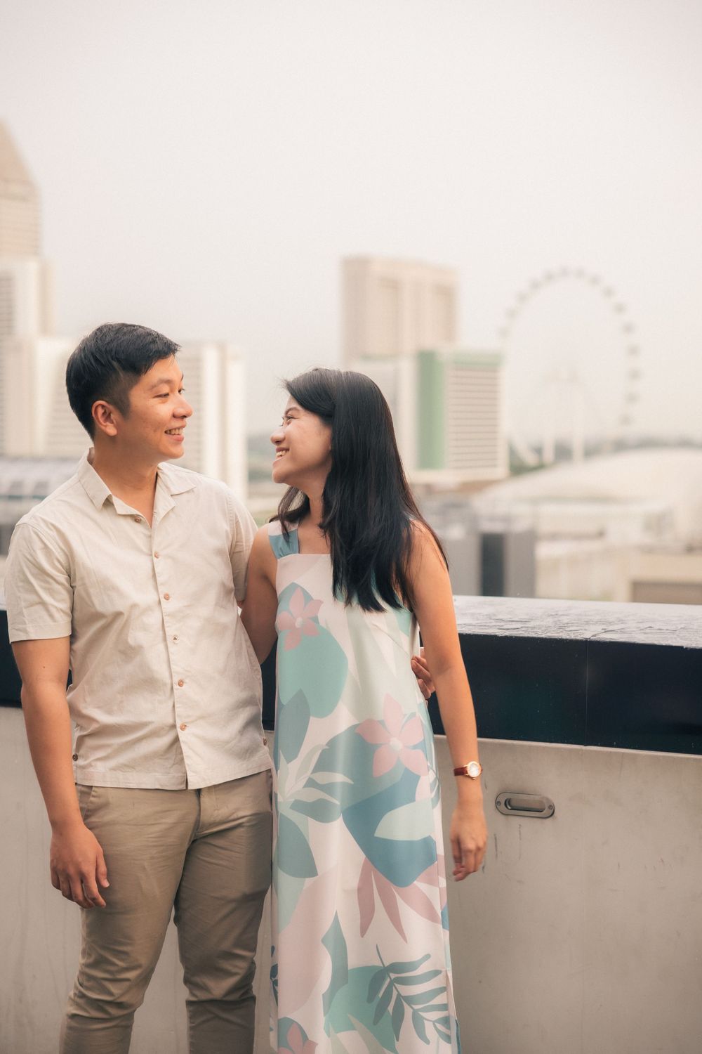 A couple shares a romantic moment on a rooftop terrace with the Singapore Flyer and city skyline visible in the background.