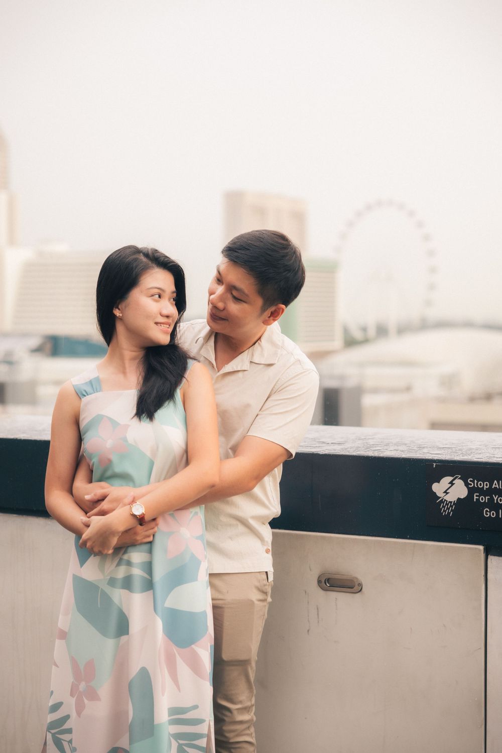 A romantic couple embraces on a rooftop with the Singapore Flyer observation wheel visible in the hazy background.