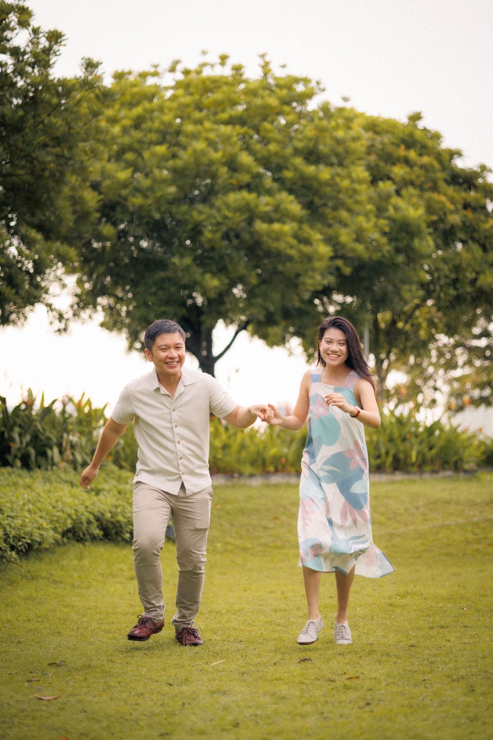 A couple holding hands and running joyfully on a grassy lawn with a leafy tree in the background.