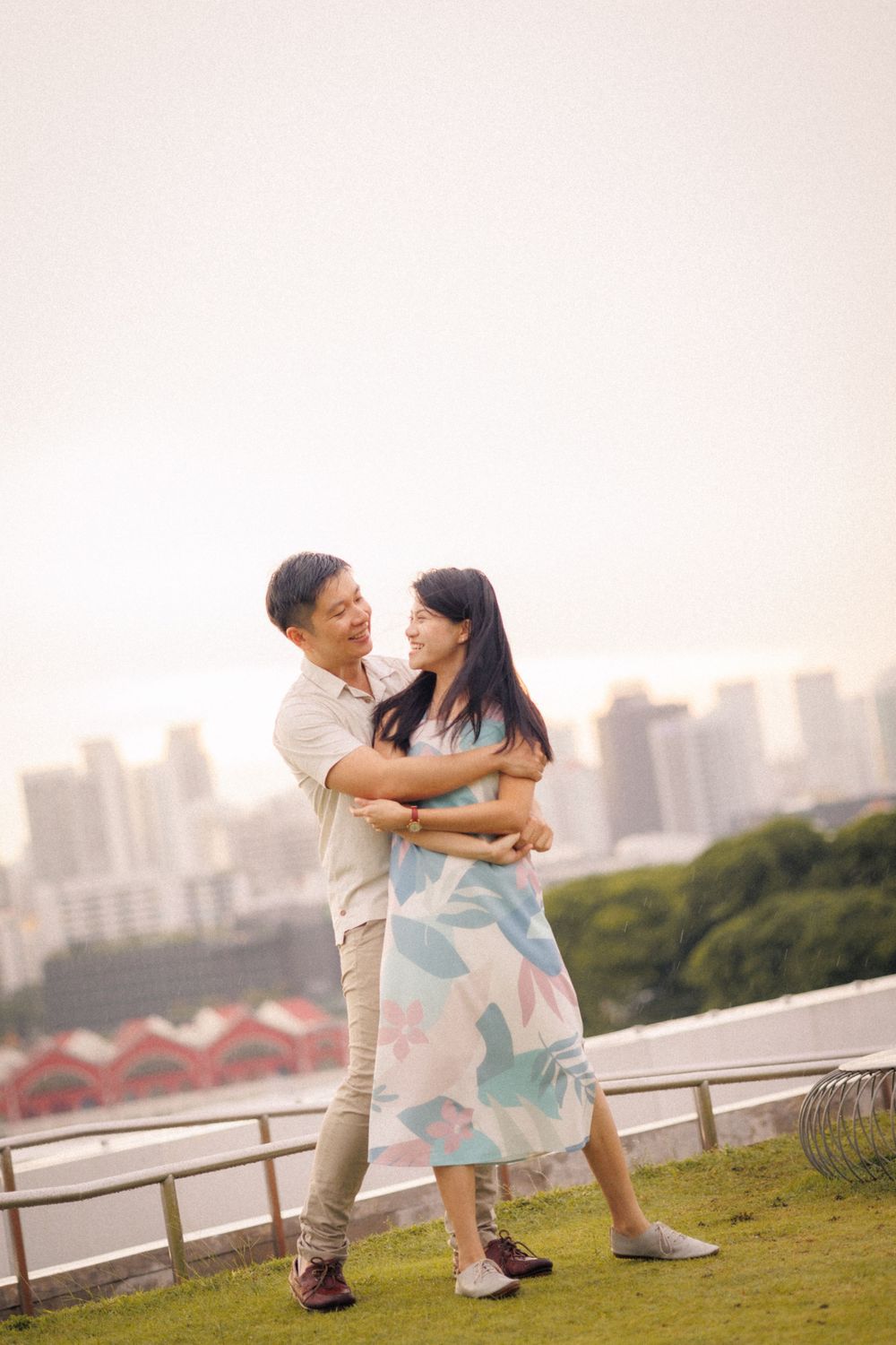 A couple embraces while standing on a grassy hill with cityscape in the background during golden hour.