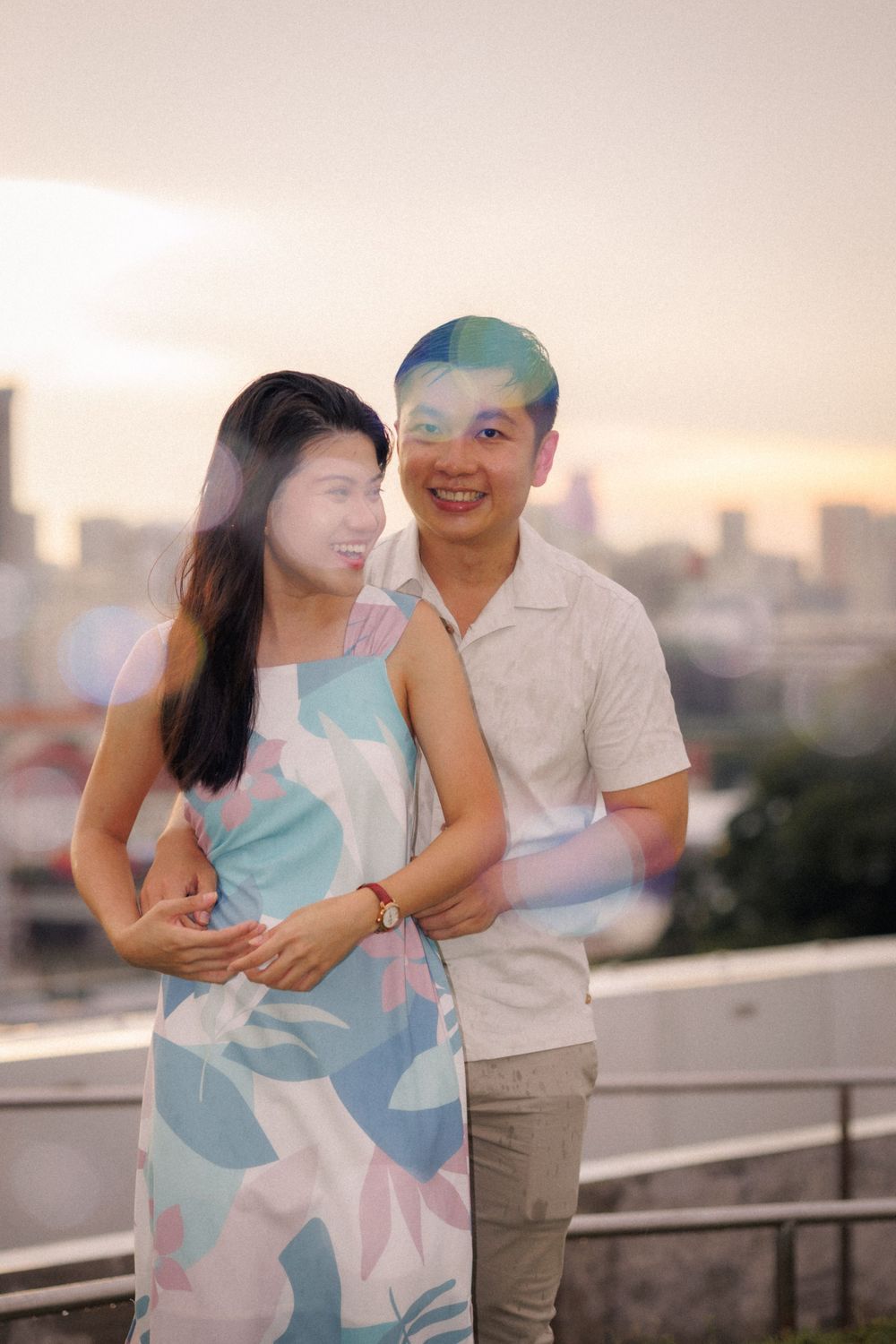 A couple shares a romantic moment on a rooftop terrace during sunset with a city skyline in the background.